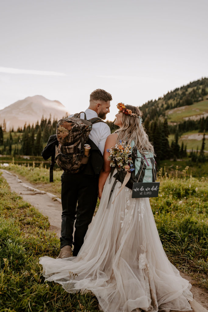 Bride and groom smile each other while their backs are to the camera as they carry hiking backpacks and prepare for a trek during their adventure elopement