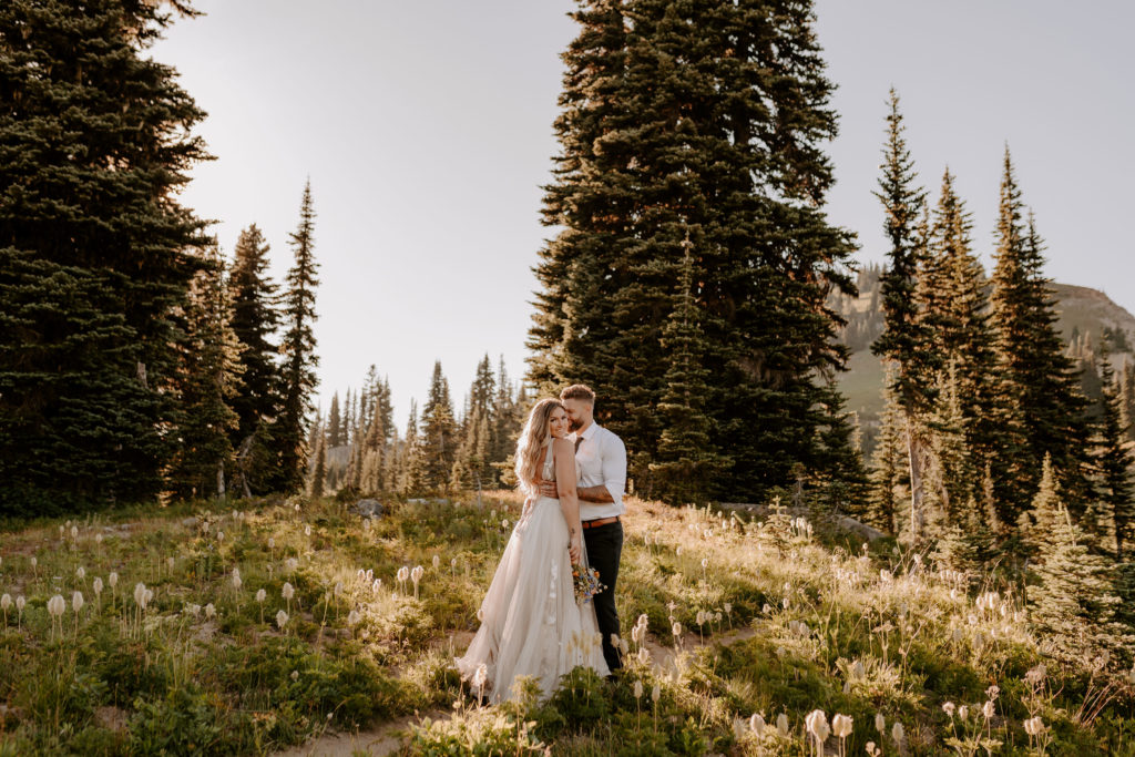 Groom embraces bride while she smiles at the camera and they stand in a field of wild flowers in the forest