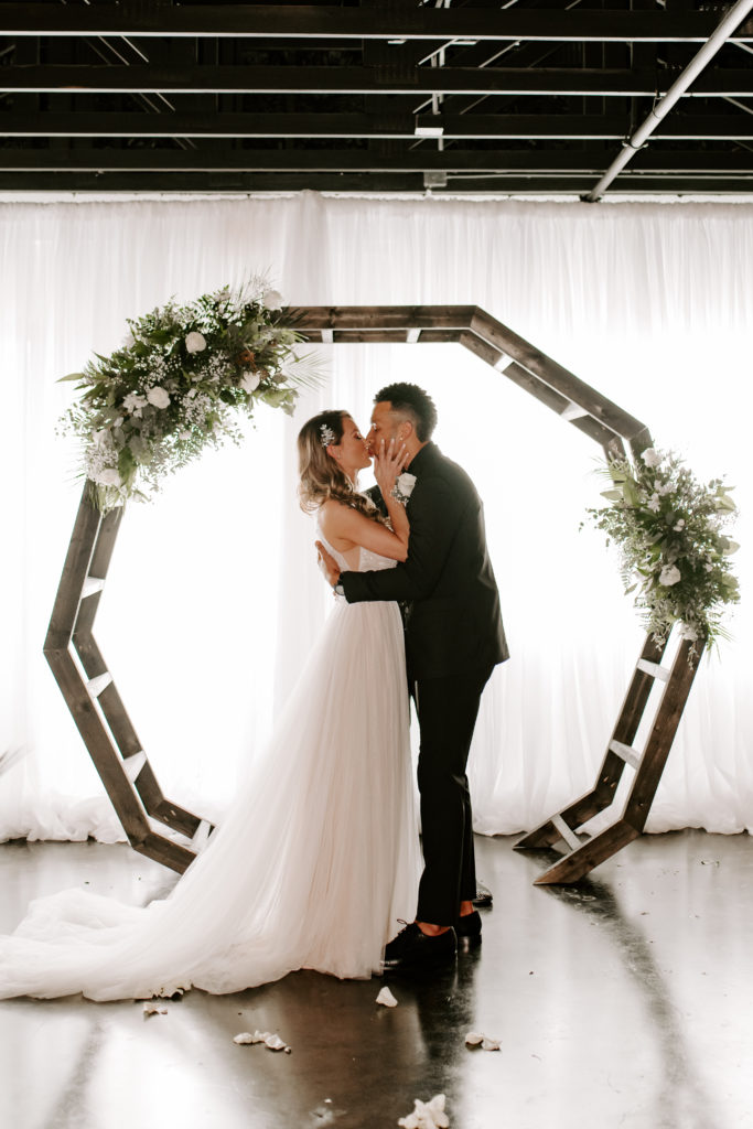 Newlywed couple sharing a kiss in front of the wedding arch at their wedding ceremony venue, captured by Off Path Photography