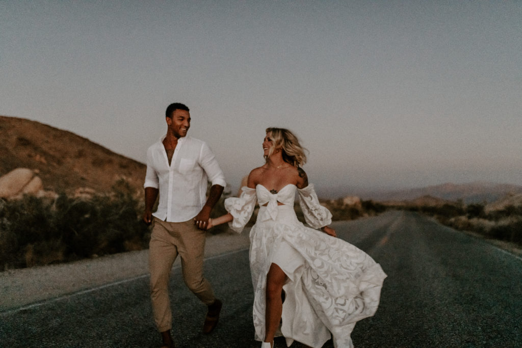 Couple running through the empty highway while holding hands and smiling at each other during their elopement shoot with Off Path Photography