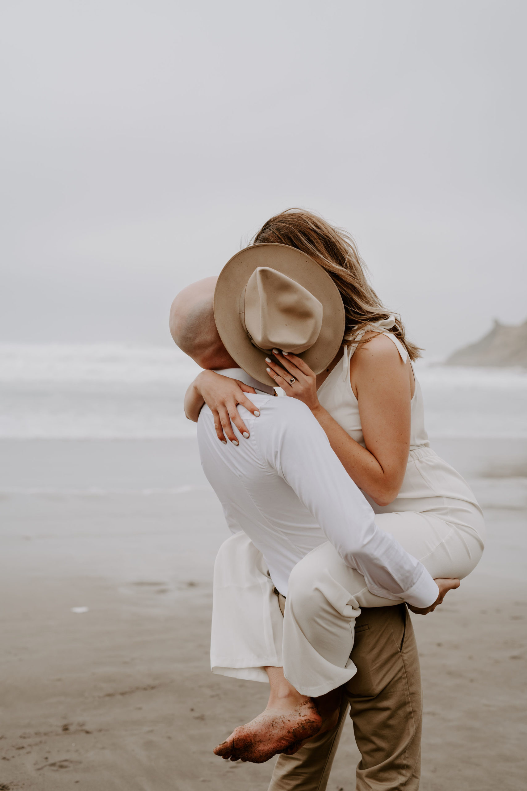 Guy carries girl as they share a kiss behind a hat at Cape Kiwanda during their elopement shoot with Off Path Photography